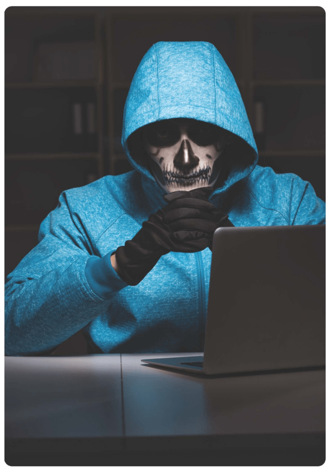 A man in a blue hoodie and skull painted on his face sitting in front of a laptop on a metal table
