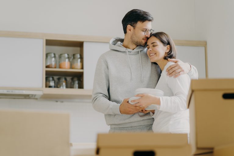 Young couple in love embrace and kiss with tender, hold pile of white plates, stand in kitchen during moving day, surrounded with many carton boxes filled with personal belongings, unpack stuff